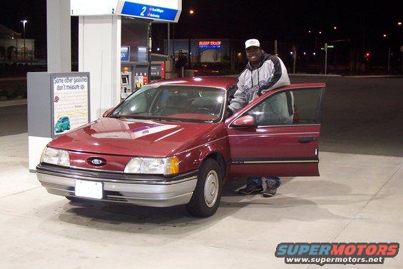 Man standing by a red 1992 Ford Taurus sedan at a gas station at night. The driver's side doors are open