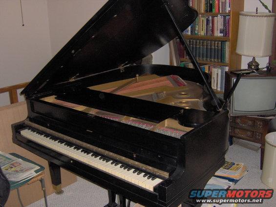 Black grand piano with lid open, showing strings and hammers, in a room with bookshelves and a television