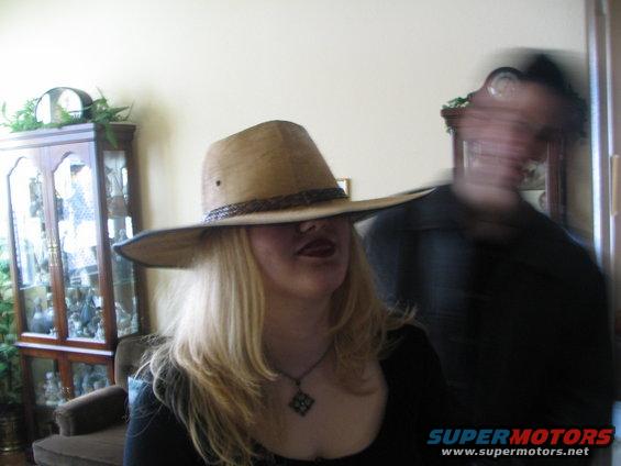 Woman in a wide-brimmed hat and necklace, with a blurry person behind her