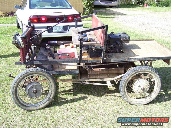 Custom flatbed vehicle with engine and wooden bed, parked behind a white sedan