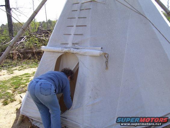 White teepee with a person entering, outdoors with trees and debris