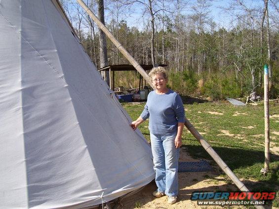 Woman in blue shirt and jeans stands by a white teepee with bamboo poles