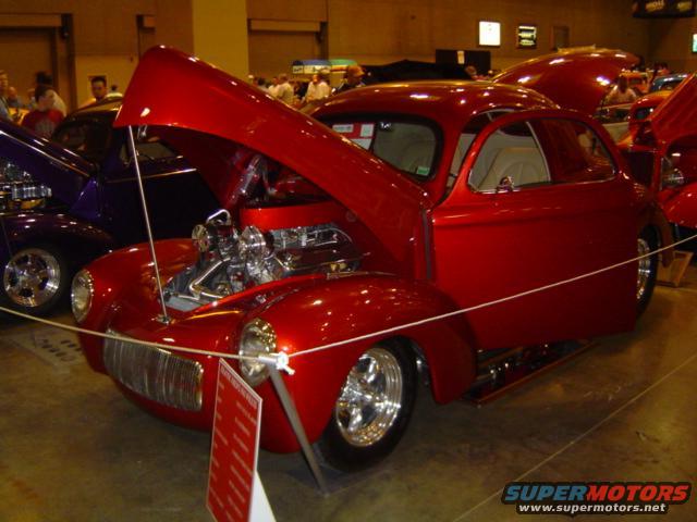 Red custom hot rod coupe with hood open, chrome engine visible. Purple hot rod in background