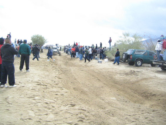 Green SUV with spare tire on rear, parked on sandy terrain with people around
