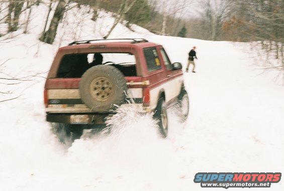 Red and white Ford Bronco with spare tire on back driving through snow