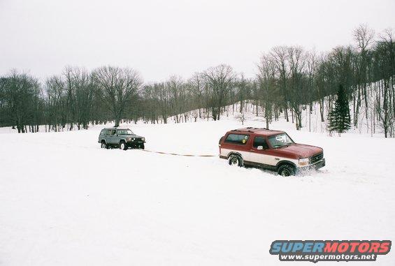 Red and white 1995 Ford Bronco towing a grey SUV in the snow