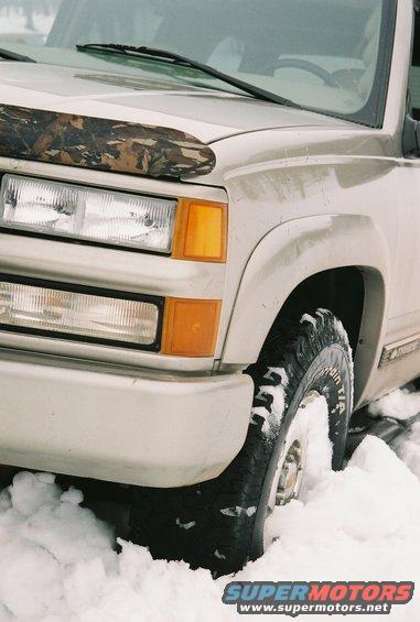 Tan pickup truck front corner in snow, camouflage bug deflector on hood, clear headlights