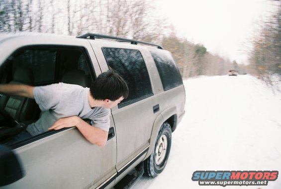 Tan 1995 Ford Bronco side view, driver leaning out window, snow on ground, trees in background
