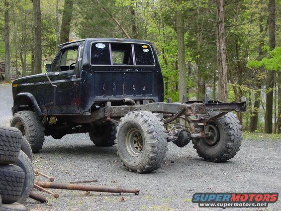 Black pickup truck chassis with large tires, missing bed and rear body panels. Visible leaf springs and differential