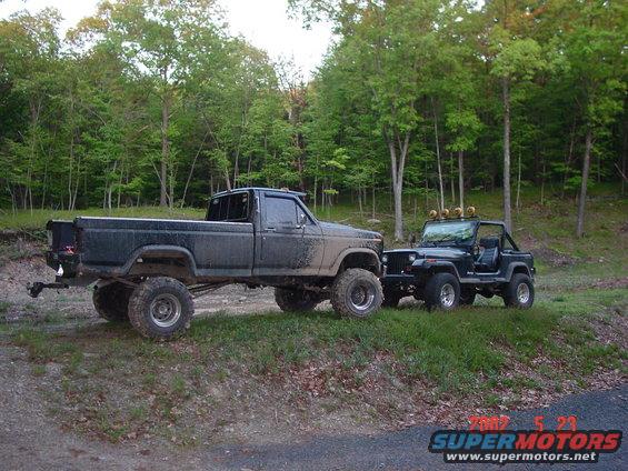 Lifted pickup truck and Jeep parked on a dirt road with trees in the background. The truck appears to be from the early 1980s