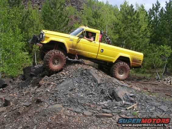 Yellow 1986 Jeep J-10 pickup truck with large off-road tires climbing a rocky incline