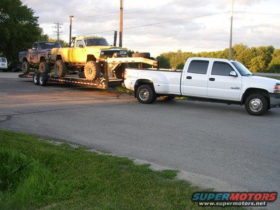 White dually pickup truck towing a flatbed trailer with two lifted trucks on it