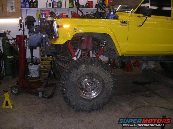 Yellow Jeep J-10 pickup truck with large tires, lifted suspension, and hood open, in a garage