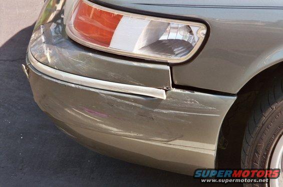 Green 2000 Mercury Grand Marquis front bumper with significant damage and scratches, right headlight visible