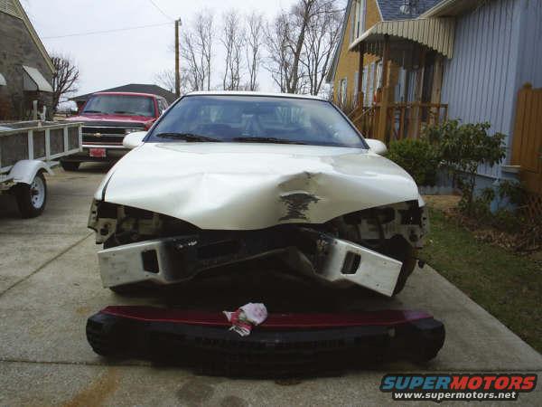 Front view of a white 1996 Ford Thunderbird with severe front-end damage. The bumper is detached and lying on the ground