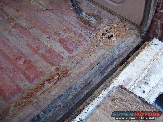 Rusted floor pan with visible holes and red paint, viewed from above