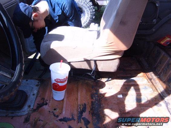 Interior of a rusty vehicle with a person leaning over the driver's seat, a Hardee's cup on the floor, and a tire visible through the window