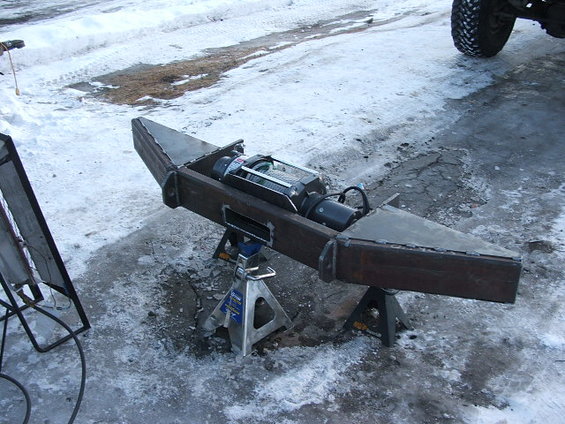 Custom steel bumper with winch mounted on jack stands, with a vehicle tire visible in the background
