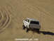 1978 Ford Bronco - A white 1978 Ford Bronco navigates sandy terrain from a rear perspective.