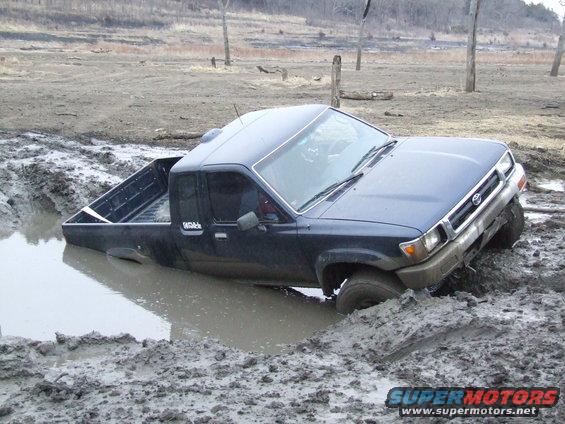 Dark blue pickup truck stuck in mud, front right wheel submerged. Visible Toyota emblem on grille