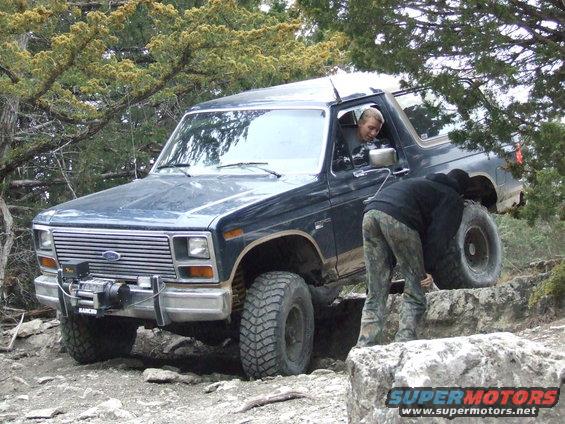 Dark blue Ford Bronco with winch on front bumper, navigating rocky terrain. Person in driver's seat, another adjusting tire