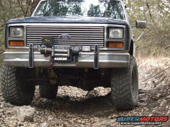 Front view of a dark blue Ford Bronco with a winch mounted on the bumper, driving on a rocky trail