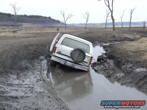 White Ford Bronco with open driver door stuck in a muddy ditch, spare tire on rear