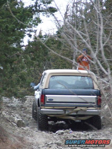 Tan and dark blue Ford Bronco on a rocky trail, rear view. A person in an orange shirt stands on top