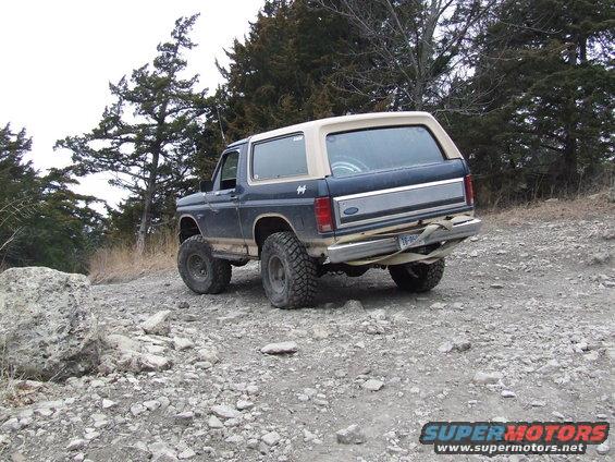 Ford Bronco on a rocky trail, rear view. It has a tan camper shell and big off-road tires