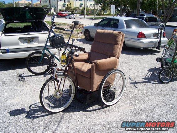 Modified wheelchair with bicycle front end and handlebars, parked next to a white car with trunk open and a silver sedan