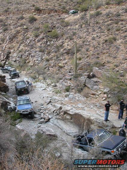 Several off-road vehicles navigating a steep, rocky trail in a desert landscape