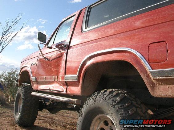 Red Ford Bronco, lifted with large off-road tires, parked on dirt. Side view shows dented door