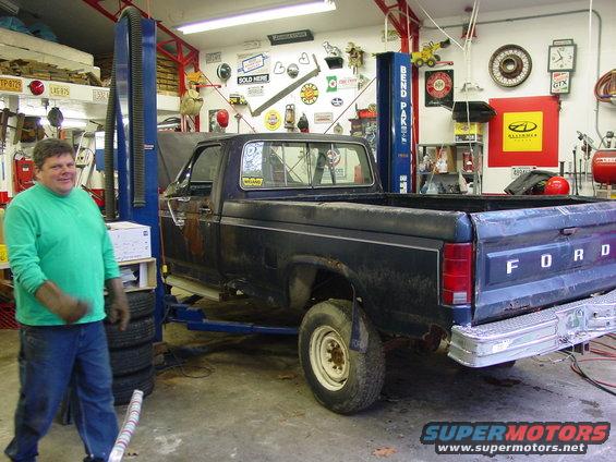 Man in green shirt stands next to dark blue Ford pickup on a lift in a garage. Truck has 'FORD' on tailgate