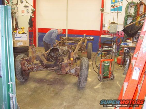 Man working on a rusty vehicle frame with engine and wheels in a garage. Welding equipment nearby