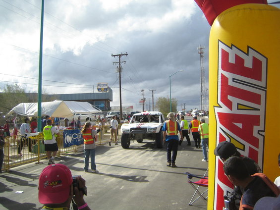 Off-road race truck with roll cage driving through an archway, people in safety vests around