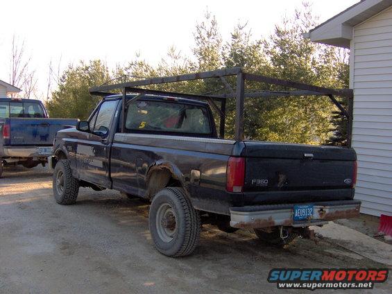 Black Ford F-350 pickup truck with a metal rack in the bed, parked next to a house. Another blue truck is visible behind it