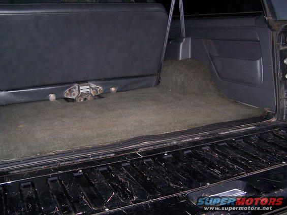Back cargo area of a vehicle with carpeted floor and a folded-down rear seat. A latch mechanism is visible on the seat base
