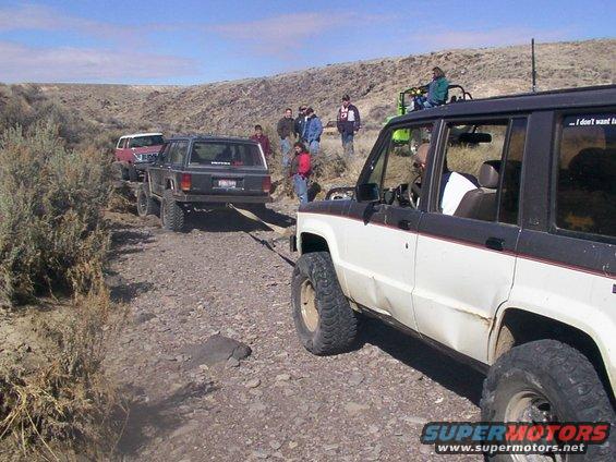 White and black SUV with dented side, towing a dark SUV on a rocky dirt road