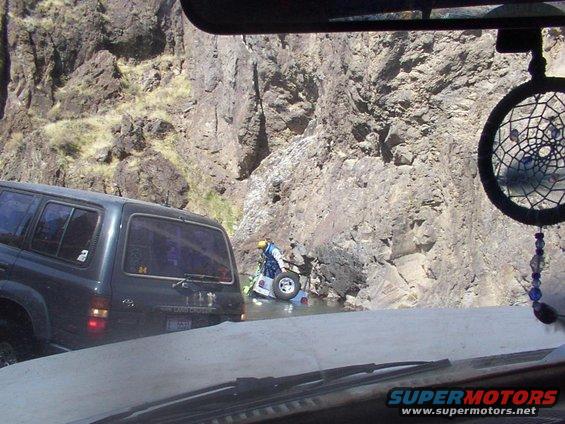 Dark SUV side view on rocky road; person near partially submerged vehicle in river