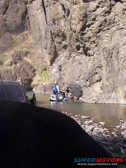 Two off-road vehicles partially submerged in a river with rocky cliffs in the background