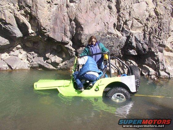 Jeep partially submerged in water with two people inside, rocky cliff face in background