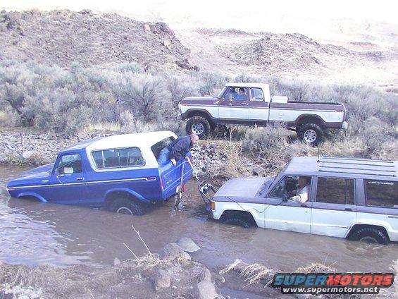 Blue 1978 Ford Bronco with white top stuck in water, being pulled by a white and grey SUV, with a brown and white pickup truck behind