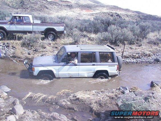 White and grey SUV being towed through a stream by a pickup truck