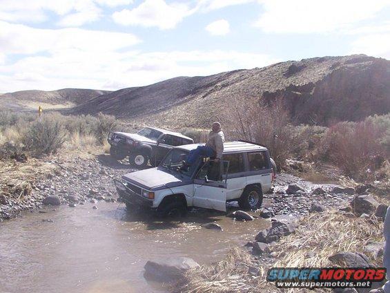 Two SUVs partially submerged in a rocky stream bed, one with a person sitting on its roof
