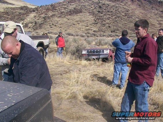 People gathered around a red truck stuck in dry brush, with another white truck nearby