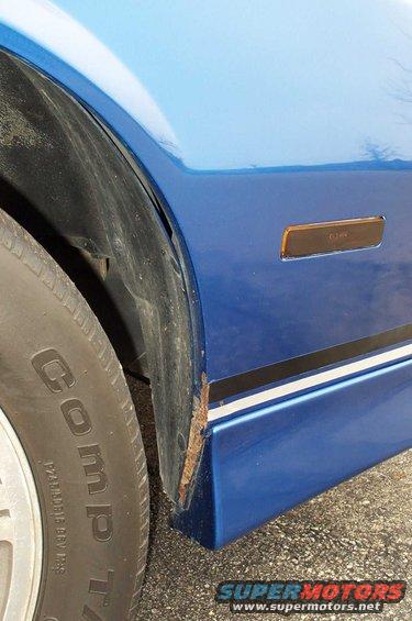 Close-up of a blue car's front fender, showing the wheel well with rust and a side marker light