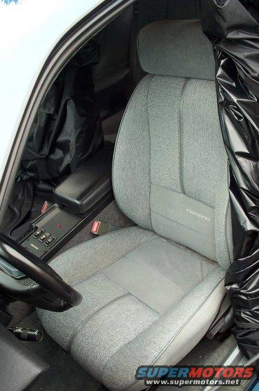 Interior shot of a 1990 Chevrolet Camaro showing the driver's seat, center console, and part of the steering wheel. The seat has 'Camaro' stitched on it