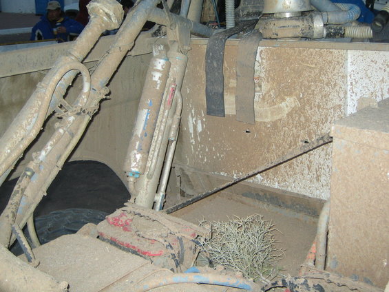 Mud-covered suspension components, shocks, and part of a tire inside a vehicle. Some dry brush is visible