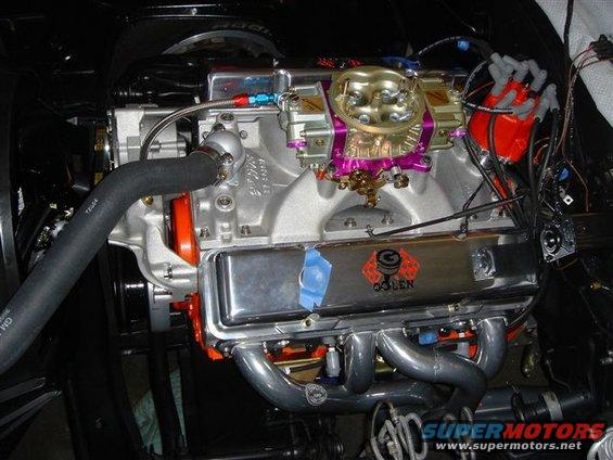Close-up of a V8 engine with a Holley carburetor, chrome valve covers, and custom headers. There's a 'QOLEN' logo on one of the valve covers