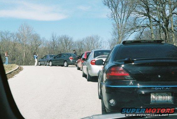 Several cars parked on a road with trees in the background. A black Pontiac is in the foreground